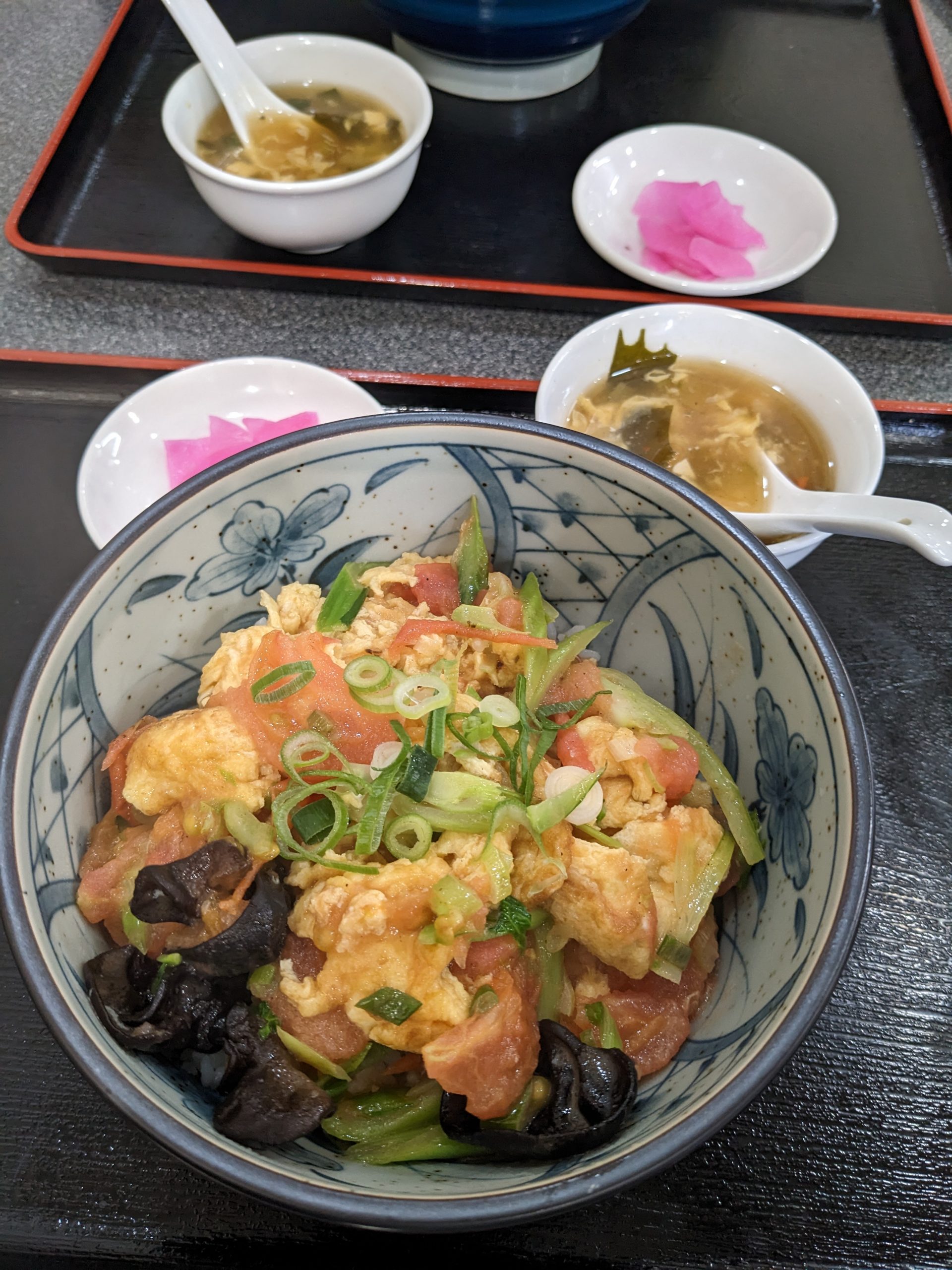 A large bowl filled with green onions, eggs, tomatoes, and mushrooms sit on a tray that has a bowl of soup and some pink pickled ginger.